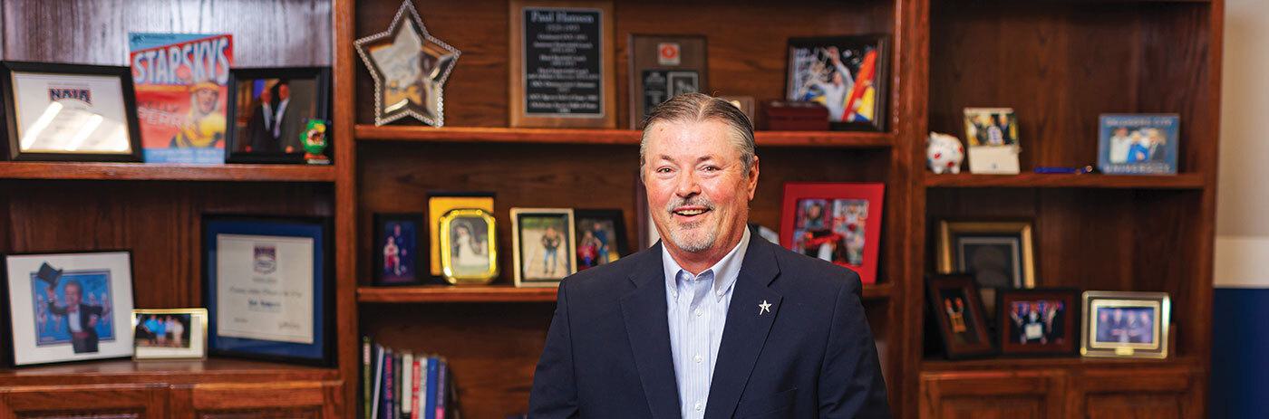 Man smiling in front of shelves of framed photos and awards